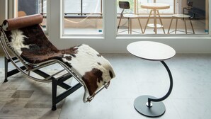 Habitación con ventanas amplias, un sillón de cuero, una mesa redonda y vistas a una calle y montañas.