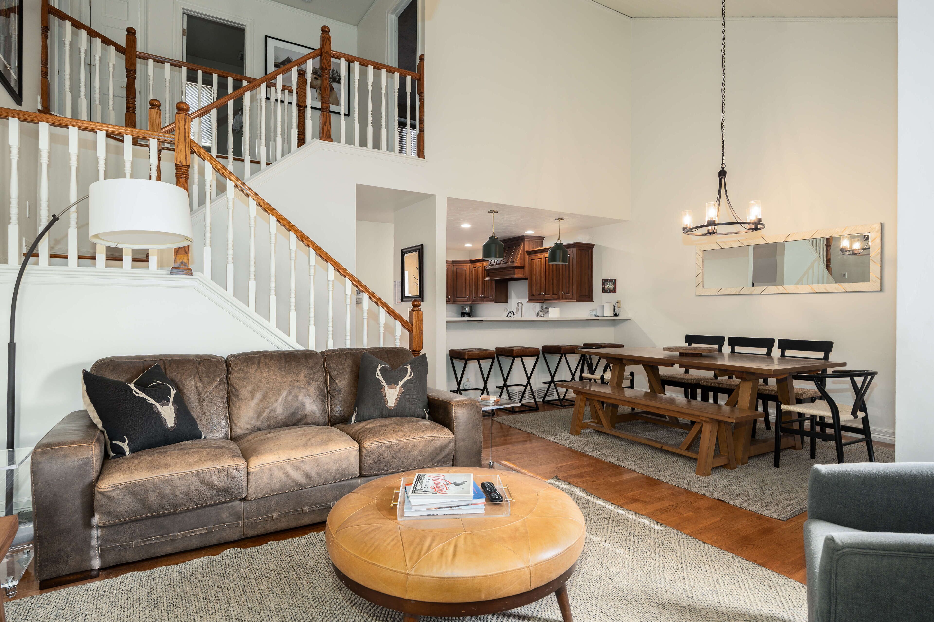 Un salón espacioso con un sofá de cuero, un otoman redondo, una mesa de comedor de madera y una escalera que conduce a un nivel superior.