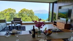 Un'area pranzo con un tavolo apparecchiato per due, un vaso di fiori e una bottiglia di vino. La stanza offre vista sul mare e sugli alberi.
