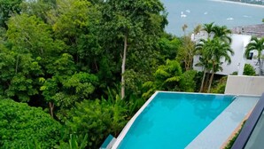 Una piscina moderna sul tetto con ringhiere in vetro, circondata da vegetazione lussureggiante e affacciata su un paesaggio costiero.