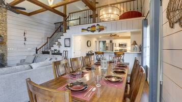 Un coin repas avec une table en bois dressée pour six personnes, entourée de chaises en bois. Un escalier avec une rampe mène à l’étage supérieur.