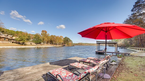 A wooden dock with lounge chairs and a red umbrella, overlooking a calm lake with trees and a dock in the background.