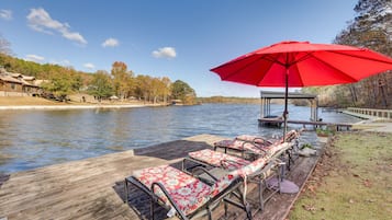 Une jetée en bois, agrémentée de chaises longues et d’un parasol rouge, surplombe un lac paisible bordé d’arbres, avec une autre jetée visible au loin.