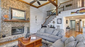 A spacious living room with a stone fireplace, a staircase, and a modern kitchen in the background.