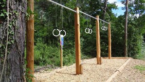 A wooden swing set with rings hanging from a horizontal bar, surrounded by trees and a gravel path. - Inter-Alp (Saint-Maurice)
