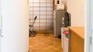 A narrow hallway with a wooden floor leading to a room with a sliding door. There are white slippers on the floor, a refrigerator, and a small table with items on it. - Dai3Himawari  401 (Hiroshima)