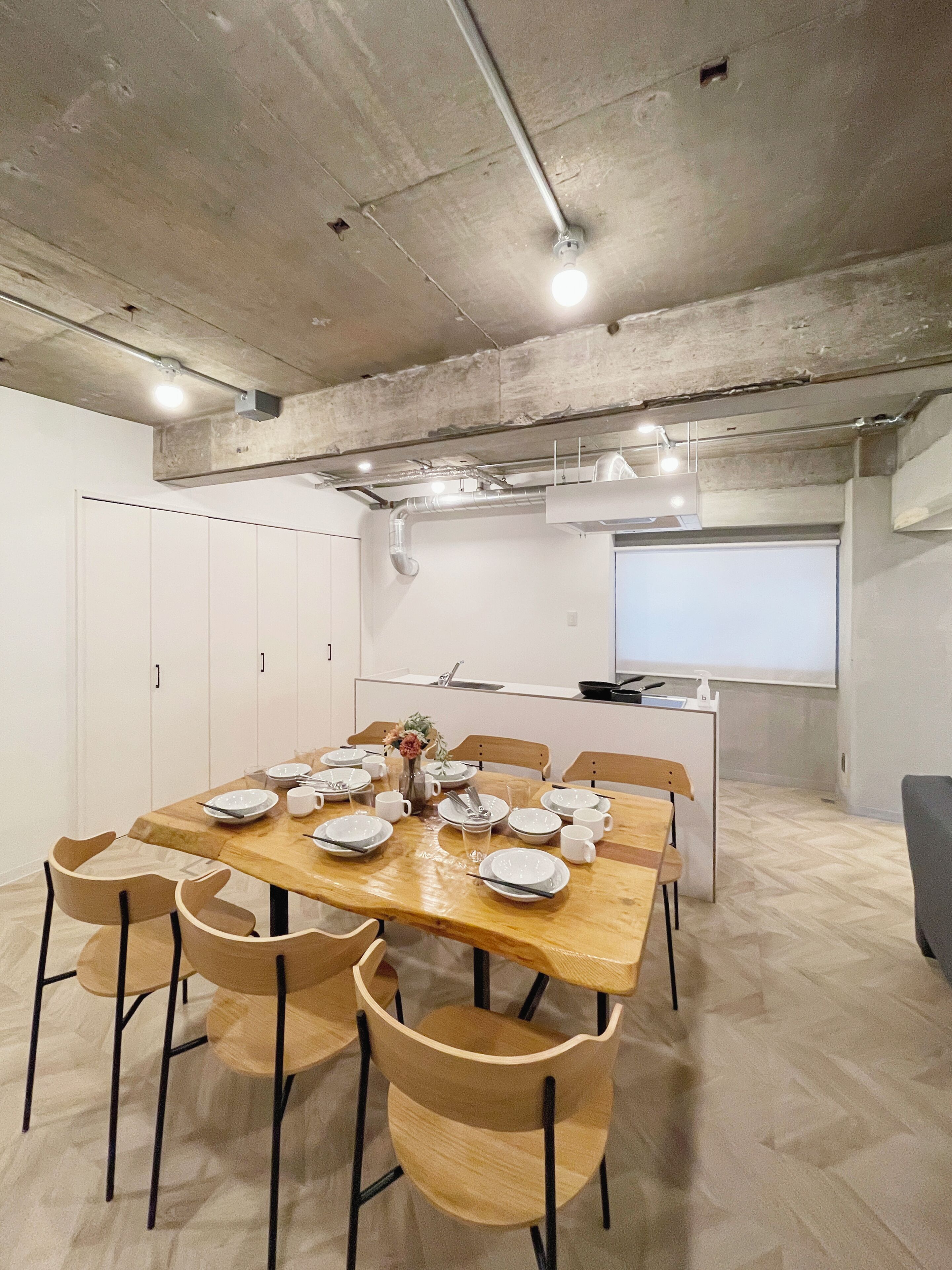 Un comedor moderno con una mesa de madera dispuesta para seis personas, rodeada de seis sillas. Se aprecia una cocina con un fregadero y una ventana al fondo.