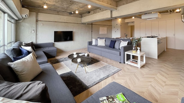 A modern living room with a grey sofa, a coffee table, and a TV. There is a kitchen area with cabinets and a sink. -  Dai3Himawari 604 (Hiroshima)