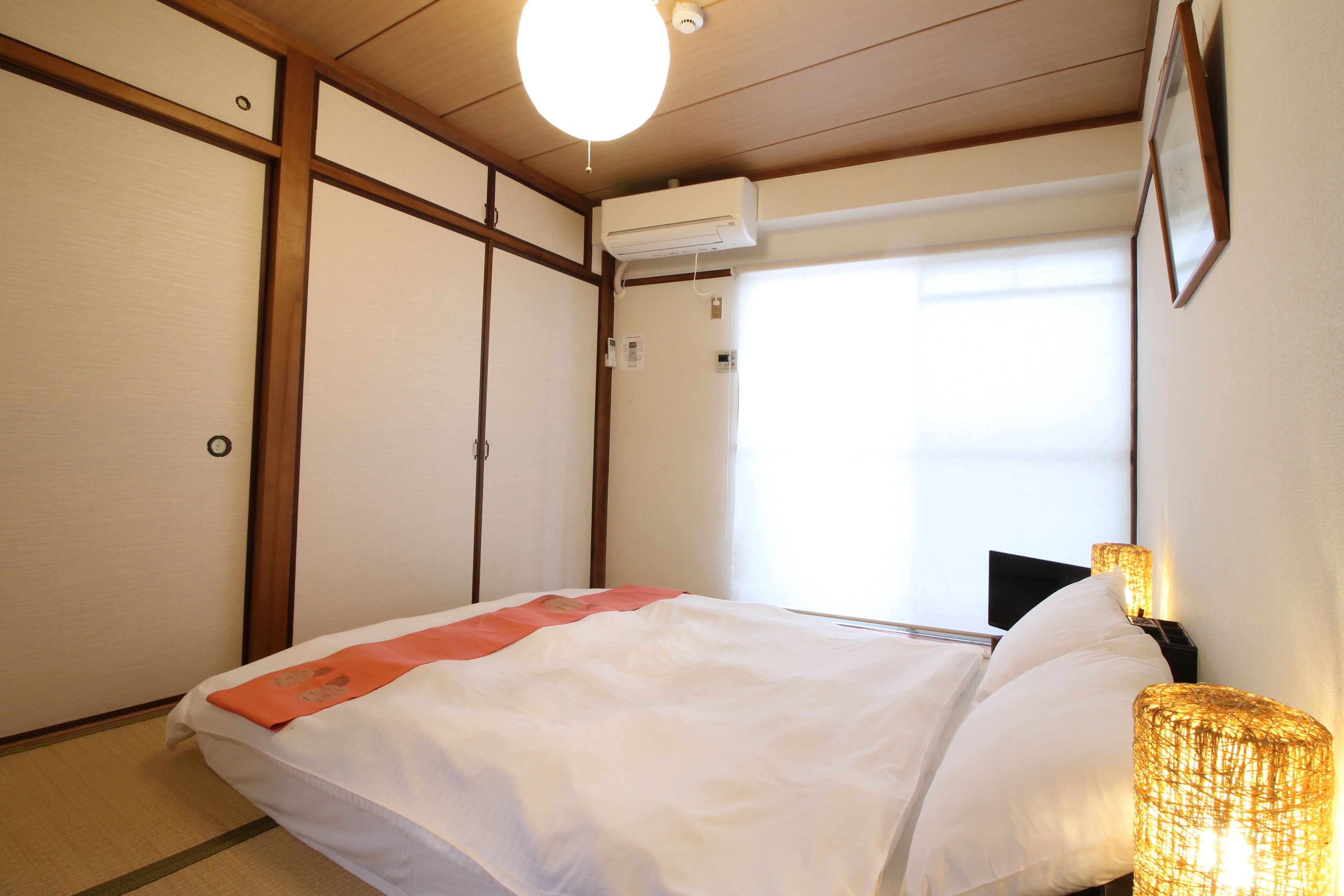 A hotel room with a bed, a bedside lamp, a window with blinds, and an air conditioning unit.