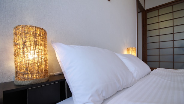 Una cama bien hecha con sábanas blancas, una mesita de noche oscura y dos lámparas de pared con pantallas tejidas.