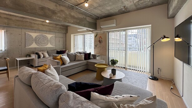 A modern living room with a sectional sofa, a small side table, and a large window with blinds. - M's Lea Building (Hiroshima)