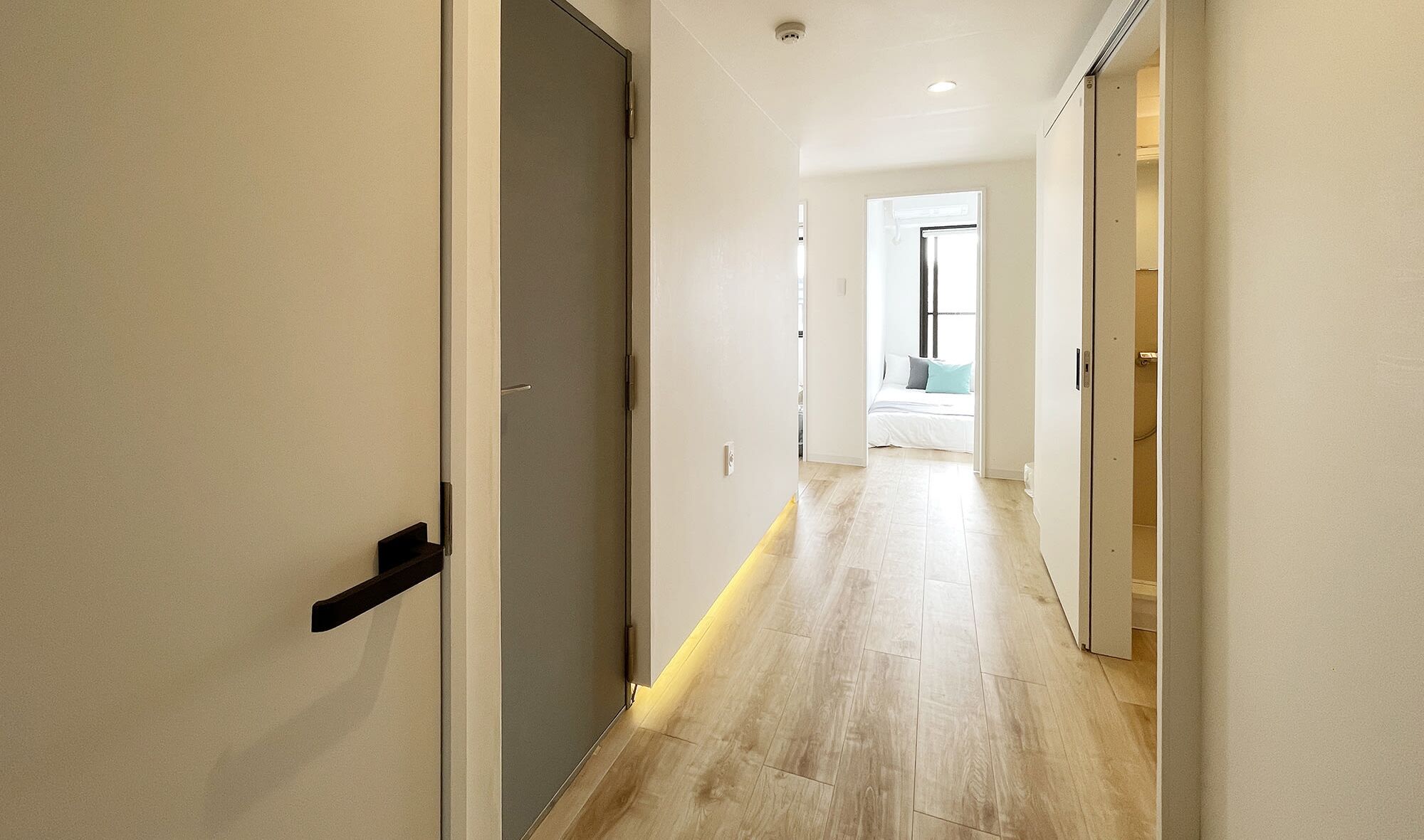 Ein moderner Flur mit Holzboden, einer Tür links und einer geöffneten Tür zu einem Schlafzimmer mit Bett und Fenster.