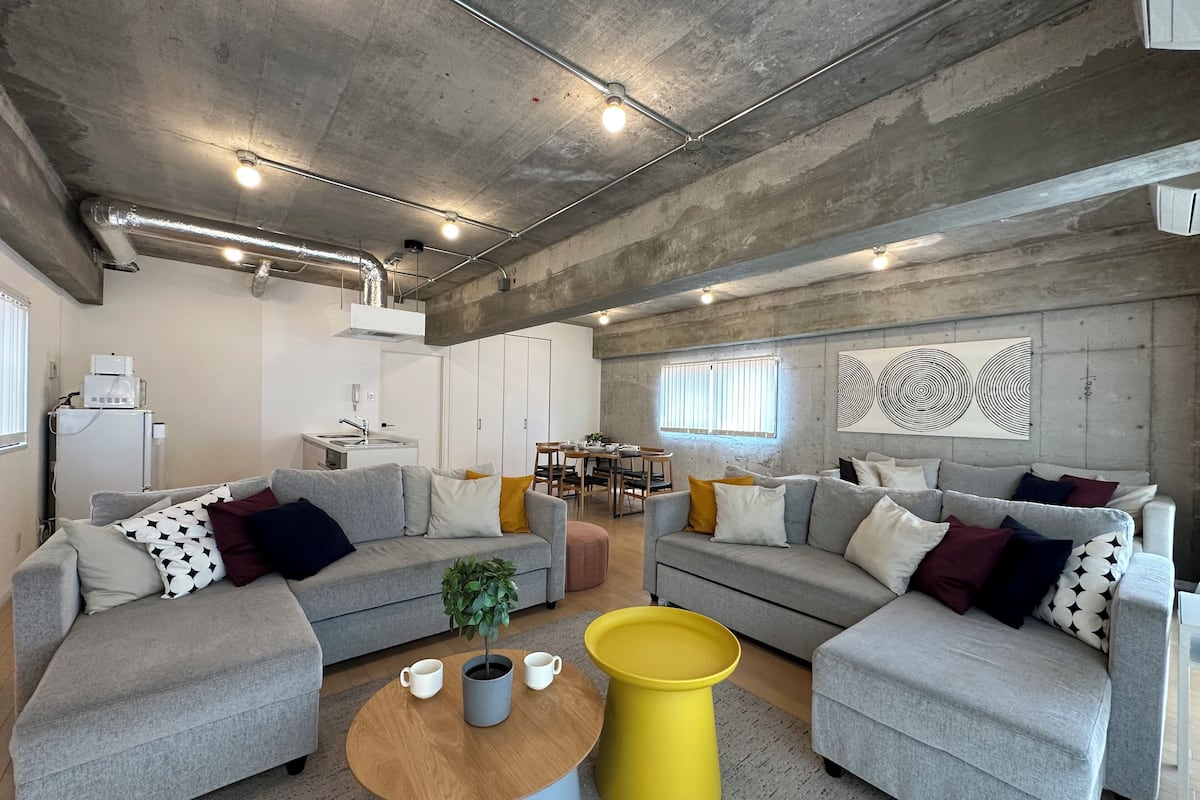 Un espacio habitable moderno con dos sofás grises, una mesa de centro de madera y una mesita auxiliar amarilla. Hay una zona de cocina con un refrigerador y un lavamanos.