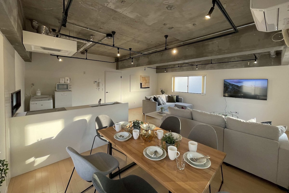 Cocina moderna con microondas, cafetera y lavabo integrados. Zona de comedor con una mesa de madera dispuesta para cuatro, rodeada de sillas grises. En el fondo, un sofá gris y un televisor.