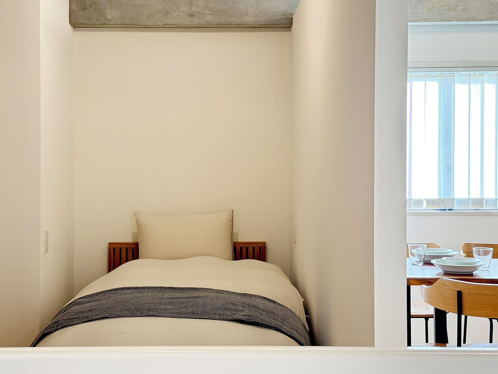 Une chambre à coucher au style minimaliste, avec un lit, une fenêtre équipée de stores et un coin repas comprenant des chaises et des assiettes.