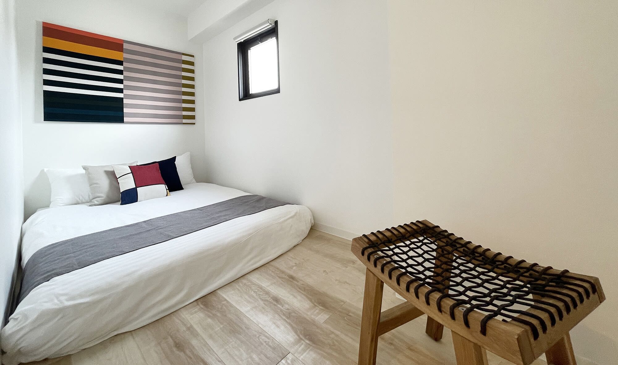 Un dormitorio con cama, almohadas, una silla de madera, una ventana y una obra de arte en la pared.