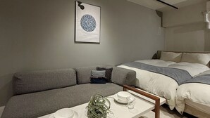 Ein modernes Schlafzimmer mit grauem Sofa, einem Bett mit weißer und grauer Bettwäsche, einem Esstisch mit Tellern und einem Glas sowie einem gerahmten Kunstwerk an der Wand.