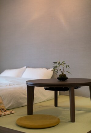 Una cama con ropa de cama blanca, una pequeña mesa redonda con una planta y un cojín amarillo.