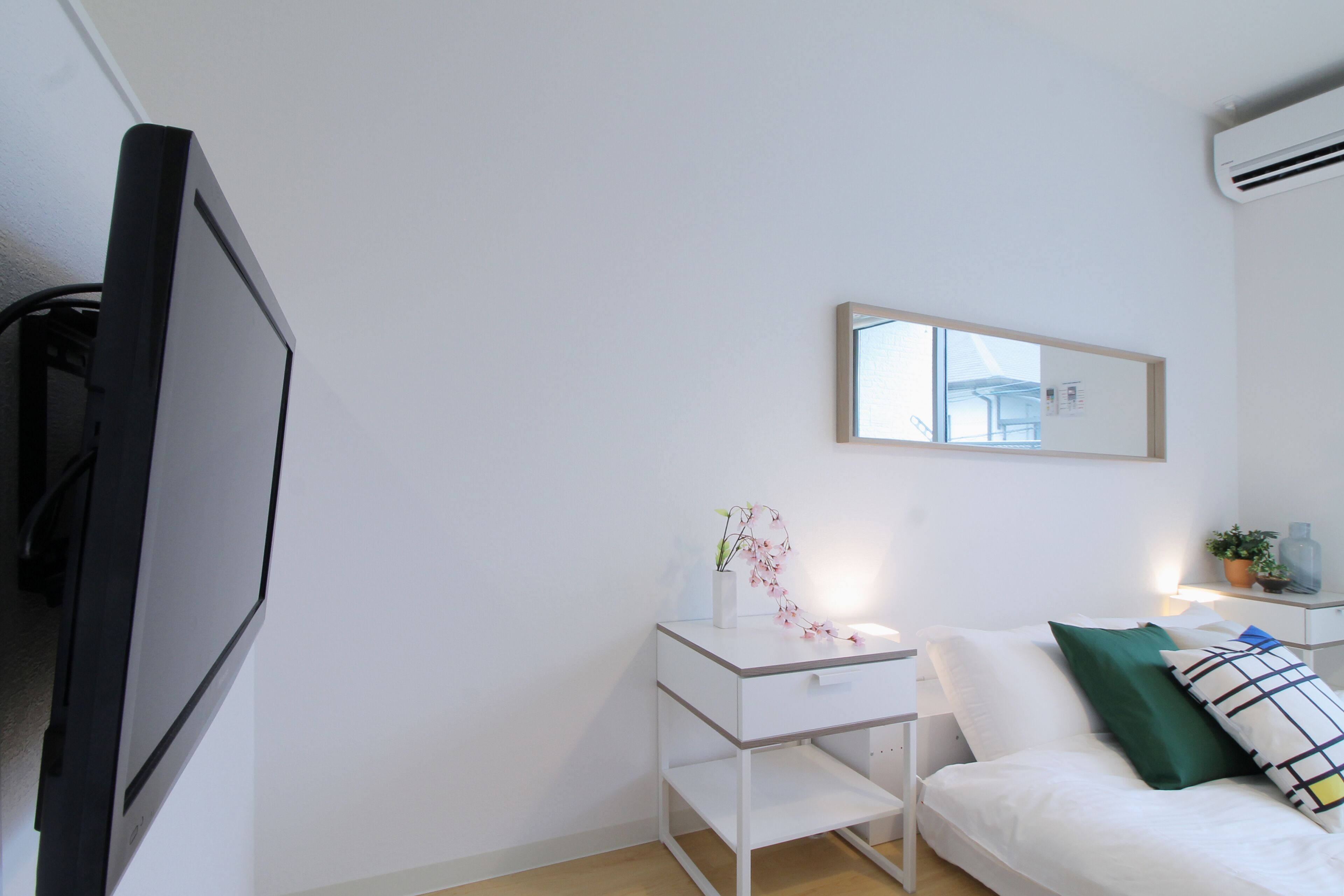 A modern hotel room with a flat-screen TV, a bedside table with a vase of flowers, a bed with pillows, and a small bedside table with a plant.