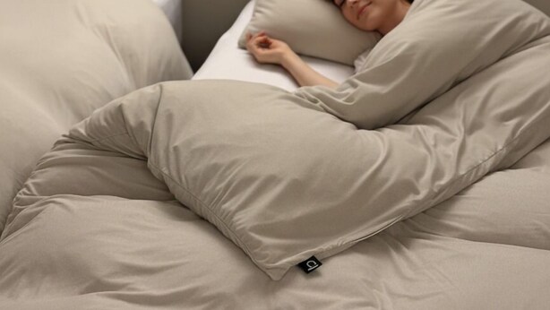 Een bed met een persoon, een beige deken, een kussen en een afstandsbediening.
