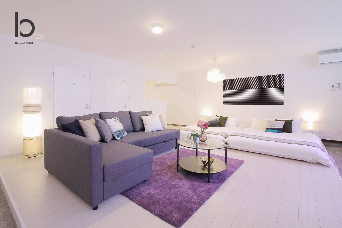 A modern hotel room with a grey sofa, a round coffee table, a white bed, and a purple rug.