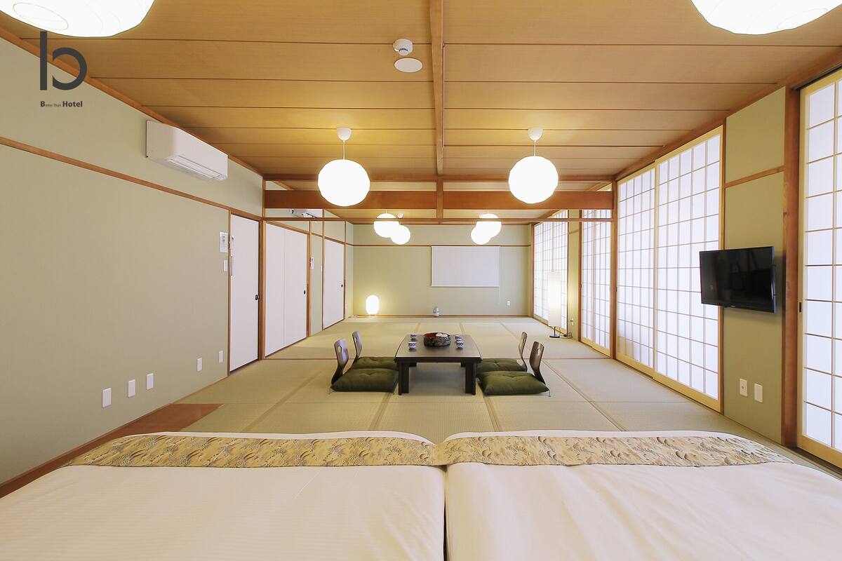 A traditional Japanese-style hotel room with tatami flooring, a tatami mat table, two tatami beds, sliding doors, and a television on the wall.