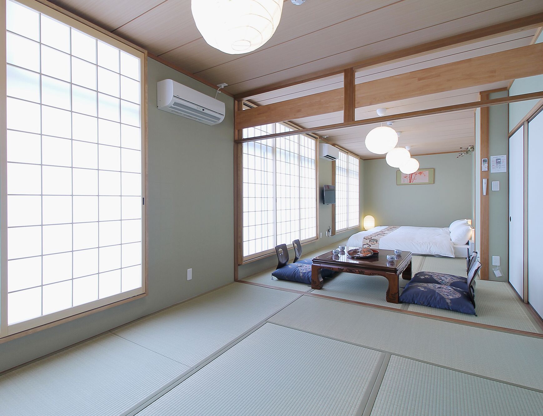 Une pièce de style japonais traditionnel, avec un sol recouvert de tatamis, un lit avec des oreillers, une petite table avec des tasses et une grande fenêtre dotée de stores à lamelles.