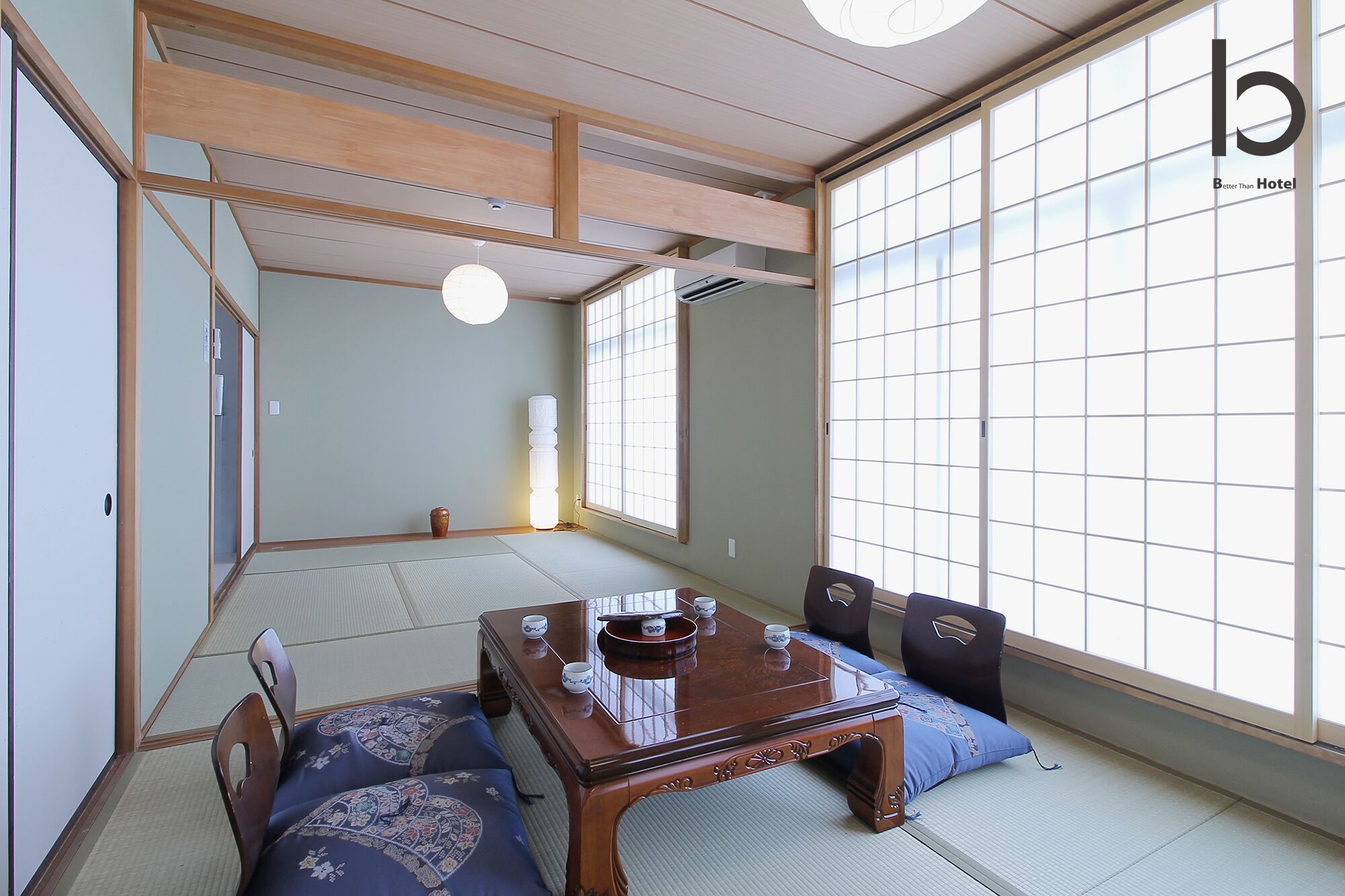 Une pièce de style japonais traditionnel, avec un sol recouvert de tatamis, une petite table en bois et un ensemble à thé, ainsi qu’une grande fenêtre à panneaux coulissants de type shoji.