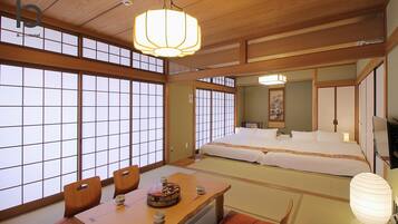 A traditional Japanese-style room with tatami flooring, a low wooden ceiling, and sliding shoji doors. There is a bed with white bedding, a table with a tea set, and a hanging lantern.