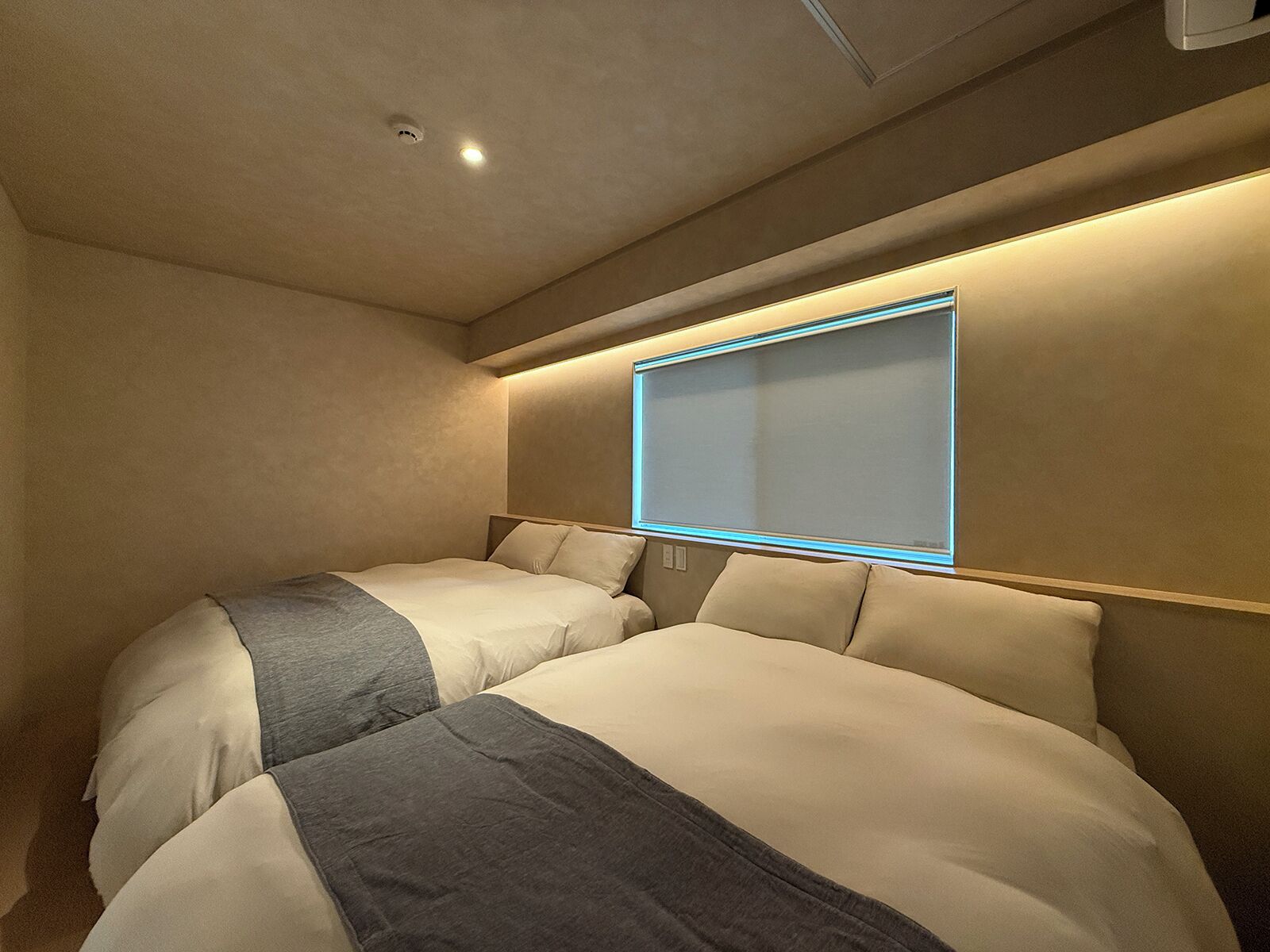 A hotel room with two beds, a large window with a blue frame, and a ceiling light.