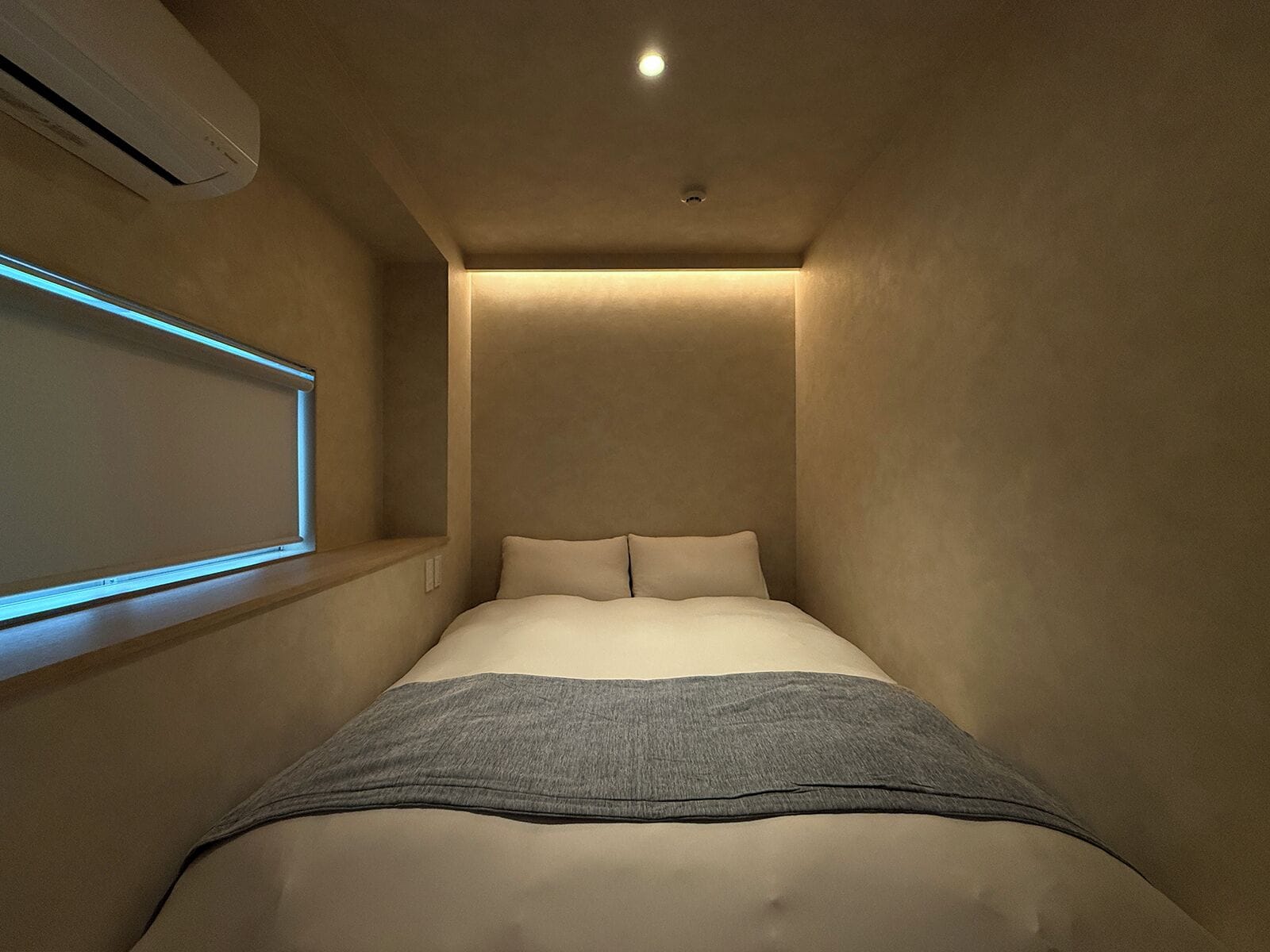 A narrow, dimly lit room with a bed, a wall-mounted projector, and a ceiling light.