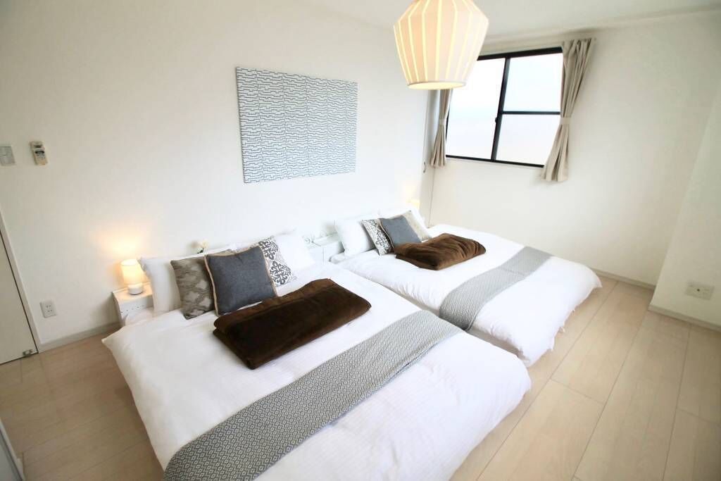 Een hotelkamer met twee bedden, een nachtkastje met een lamp, een raam met gordijnen en een kunstwerk aan de muur.