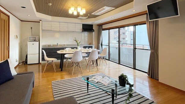 A modern living room with a sofa, a dining area with chairs, a kitchen, and a TV mounted on the wall. - Mbuilding (Hiroshima)