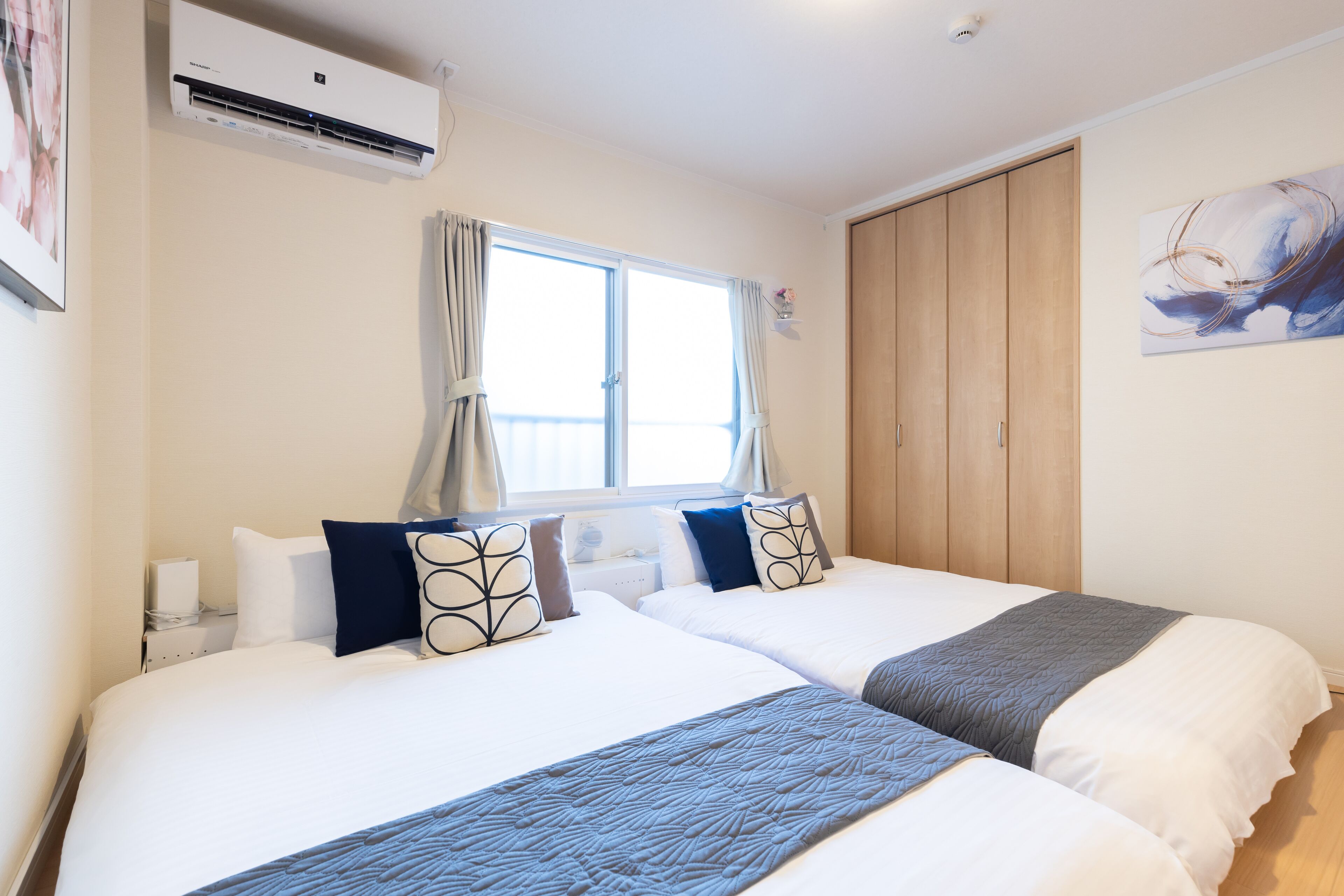 Habitación de hotel con dos camas, una ventana con cortinas, un aire acondicionado, un armario de madera y un cuadro en la pared.