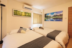 A hotel room with two beds, a window with curtains, an air conditioning unit, and two framed landscape paintings. - Sakamoto- House (Hiroshima)