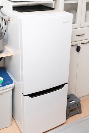 Uma geladeira Hisense branca e compacta em uma cozinha com piso de madeira, um armário branco com puxador preto e uma caixa de armazenamento azul com etiqueta verde.