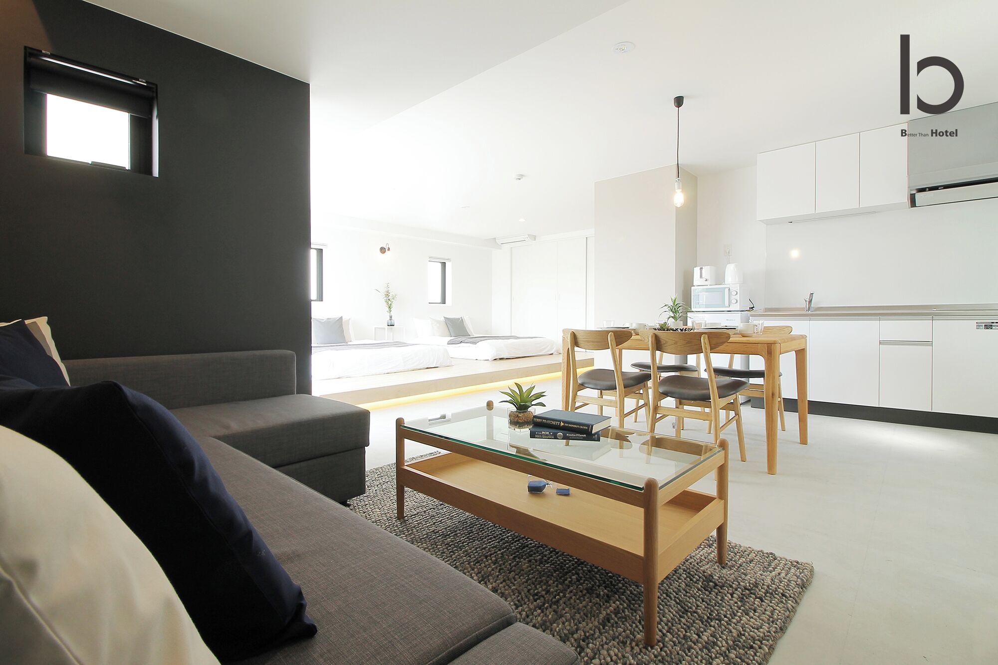 A modern hotel room with a sofa, a glass coffee table, a dining area with wooden chairs, and a kitchenette.