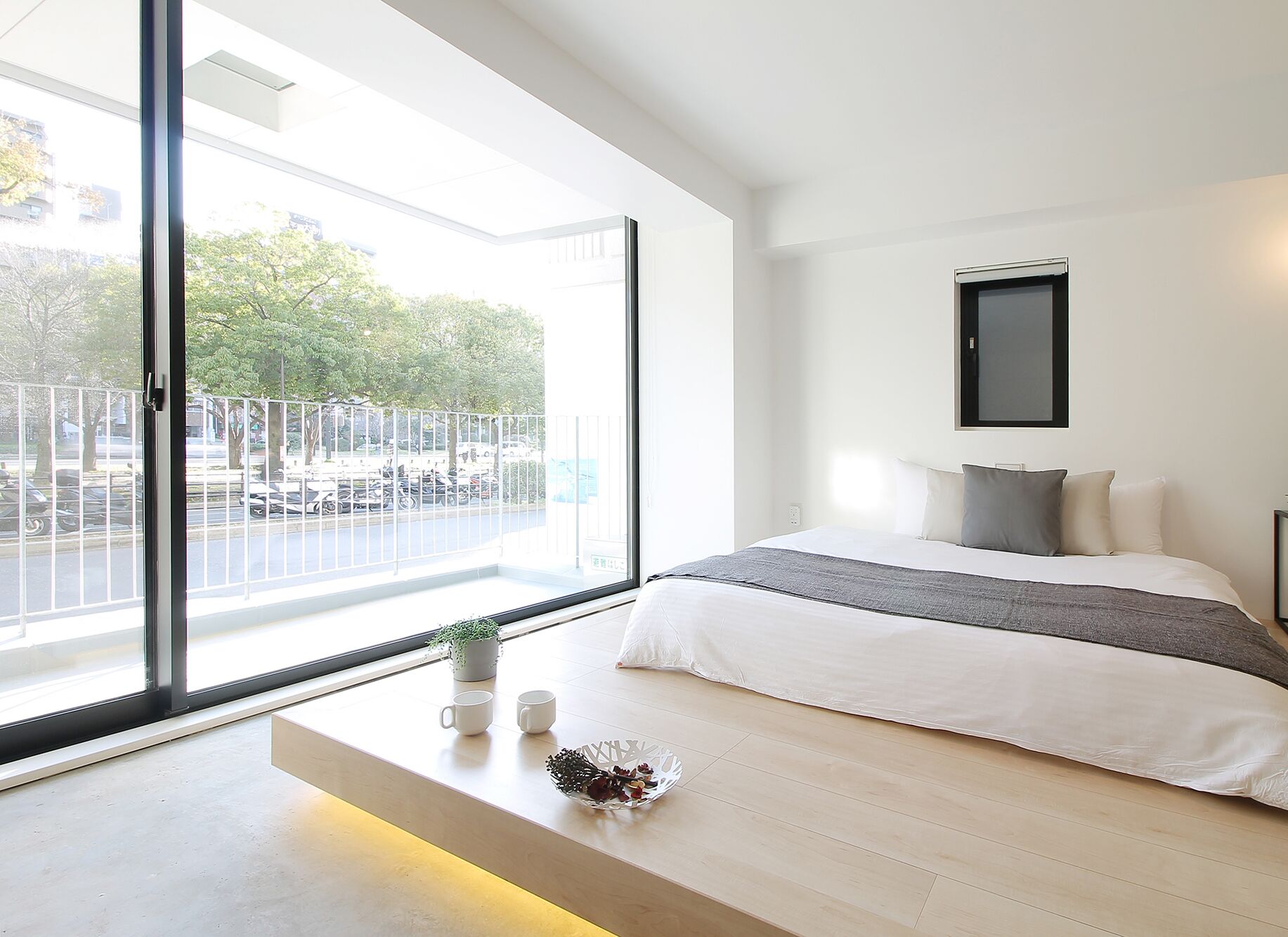 Ein modernes Schlafzimmer mit einem großen Fenster, einem Bett mit weißer und grauer Bettwäsche, einem kleinen Nachttisch mit zwei weißen Tassen und einer Topfpflanze.