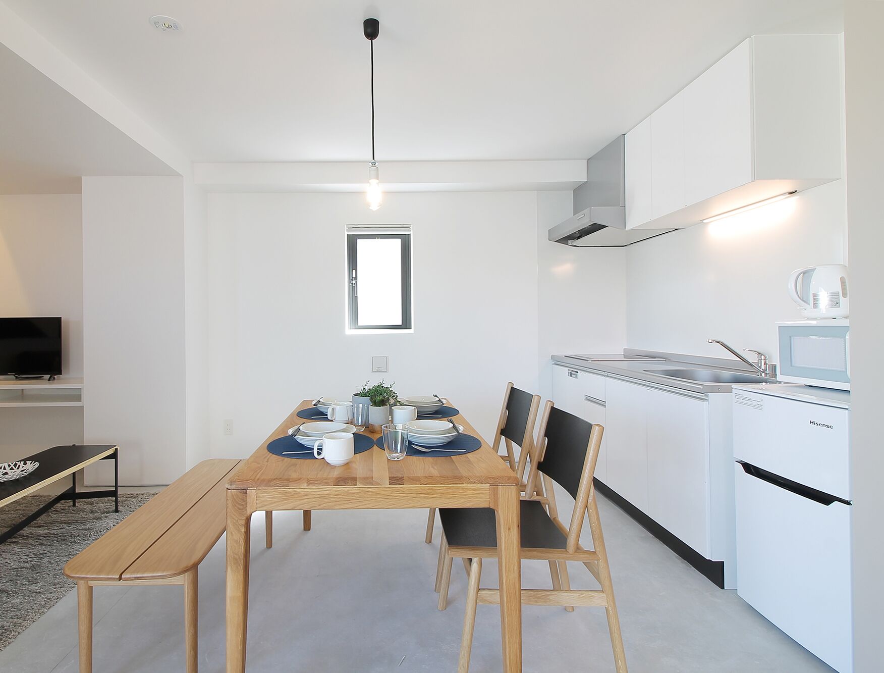 A modern kitchen with white cabinets, a built-in stove, and a microwave. There is a dining table with chairs and a bench, and a small window above the table.