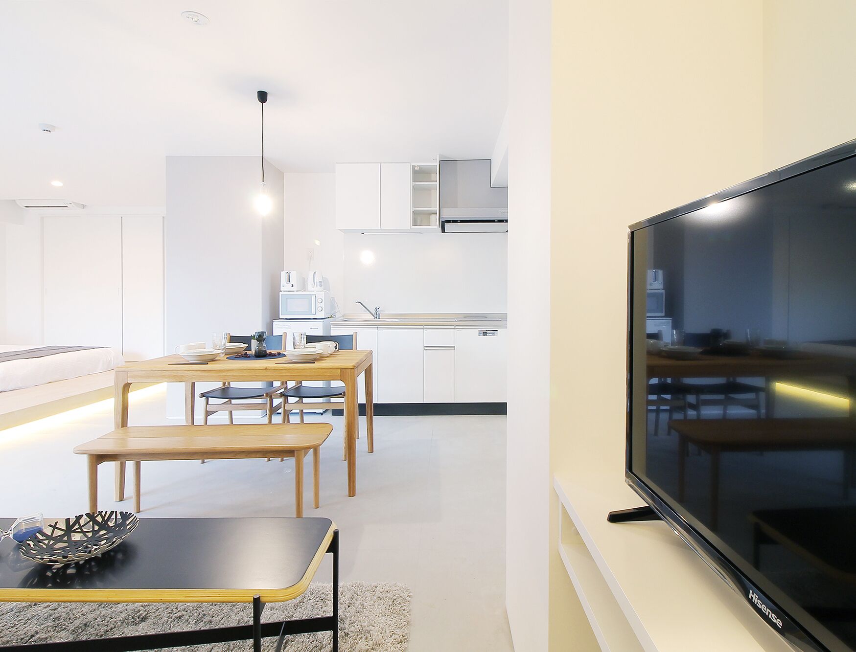 A modern kitchen with white cabinets, a dining area with wooden tables and chairs, and a large flat-screen TV mounted on the wall.