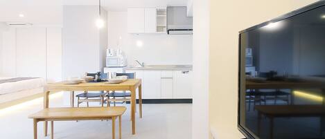 A modern kitchen with white cabinets, a dining area with wooden tables and chairs, and a large flat-screen TV mounted on the wall.