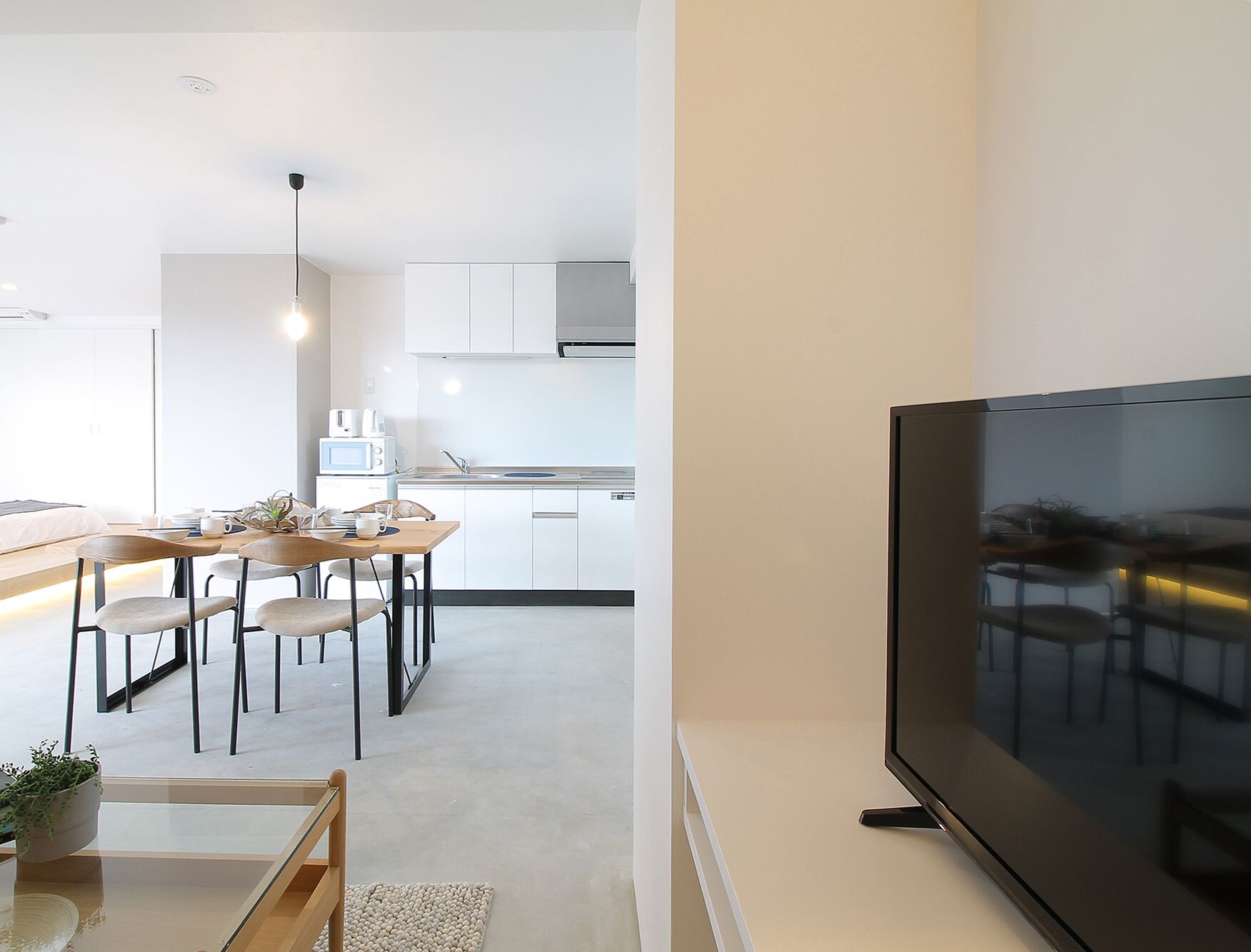 A modern kitchen with white cabinets, a dining area with wooden chairs, and a large flat-screen TV mounted on the wall.