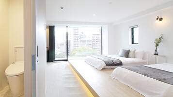 A modern bedroom with a large bed, a bedside table, a window with a view of buildings, and a toilet.