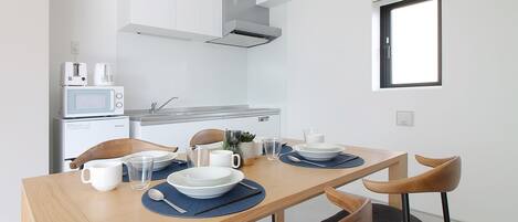 Een kleine keuken met een magnetron, broodrooster en spoelbak. Een eettafel met stoelen en een raam erboven.
