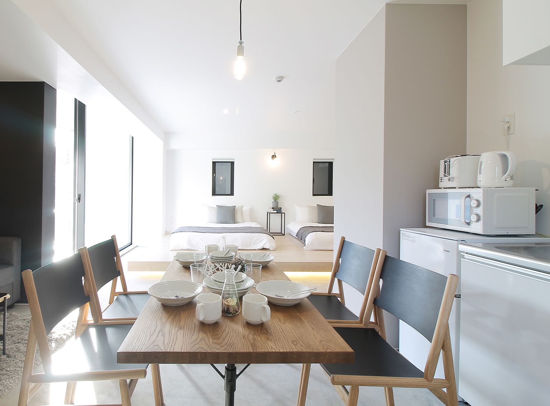 A modern kitchen with white appliances, a dining area with a wooden table and chairs, and a view of a bedroom with a bed and bedside tables.