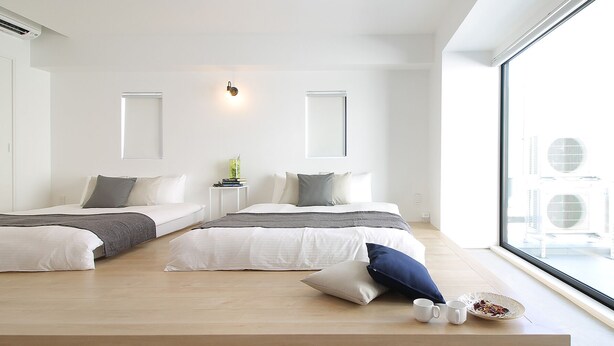 A modern hotel room with two beds, a small bedside table, a wall-mounted light, and a large window with air conditioning units visible outside.