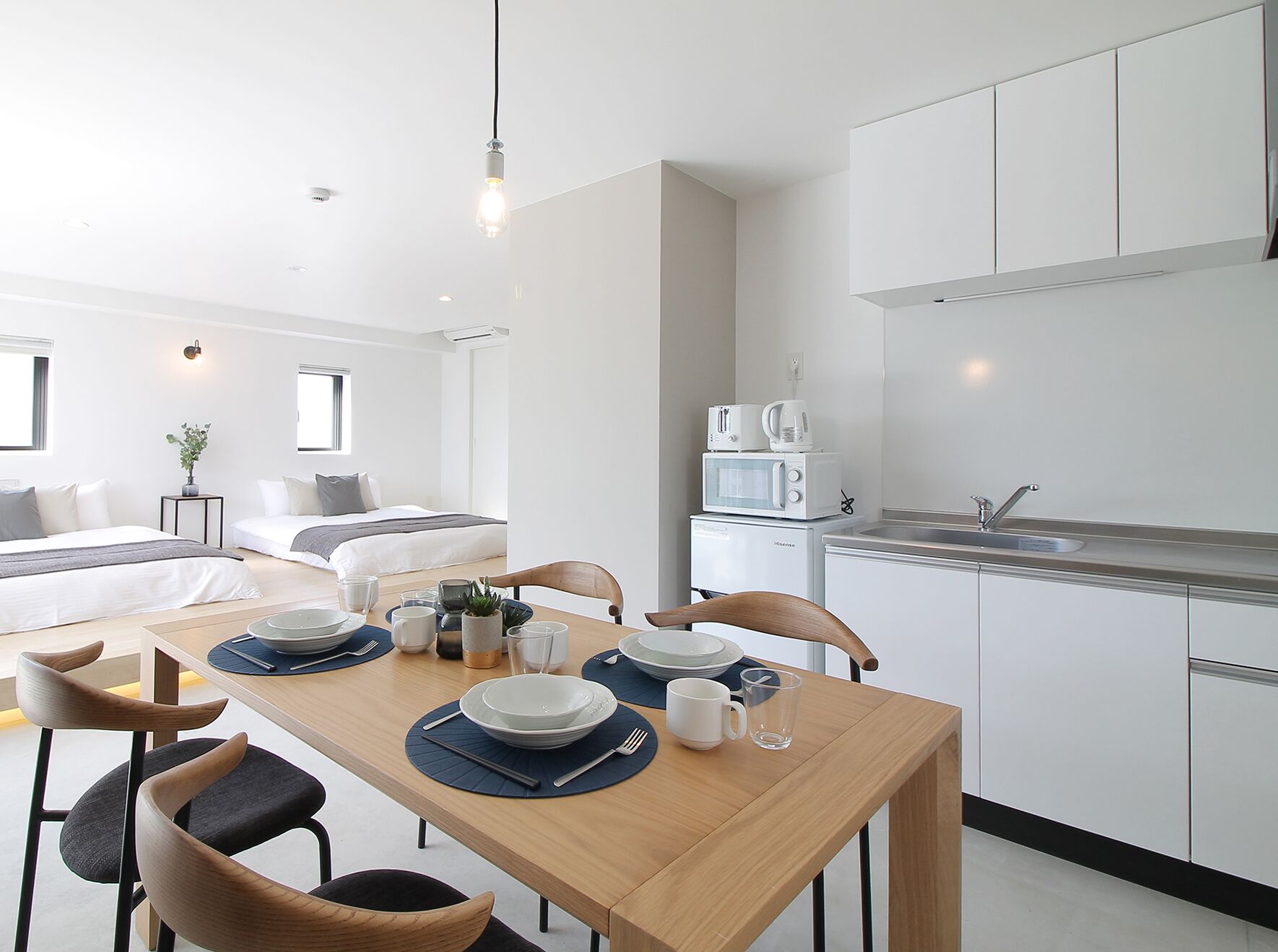 Una habitación moderna y luminosa con una mesa de comedor preparada para dos personas, una kitchenette con microondas y cafetera, una cama con cabecera gris y una pequeña planta en maceta.