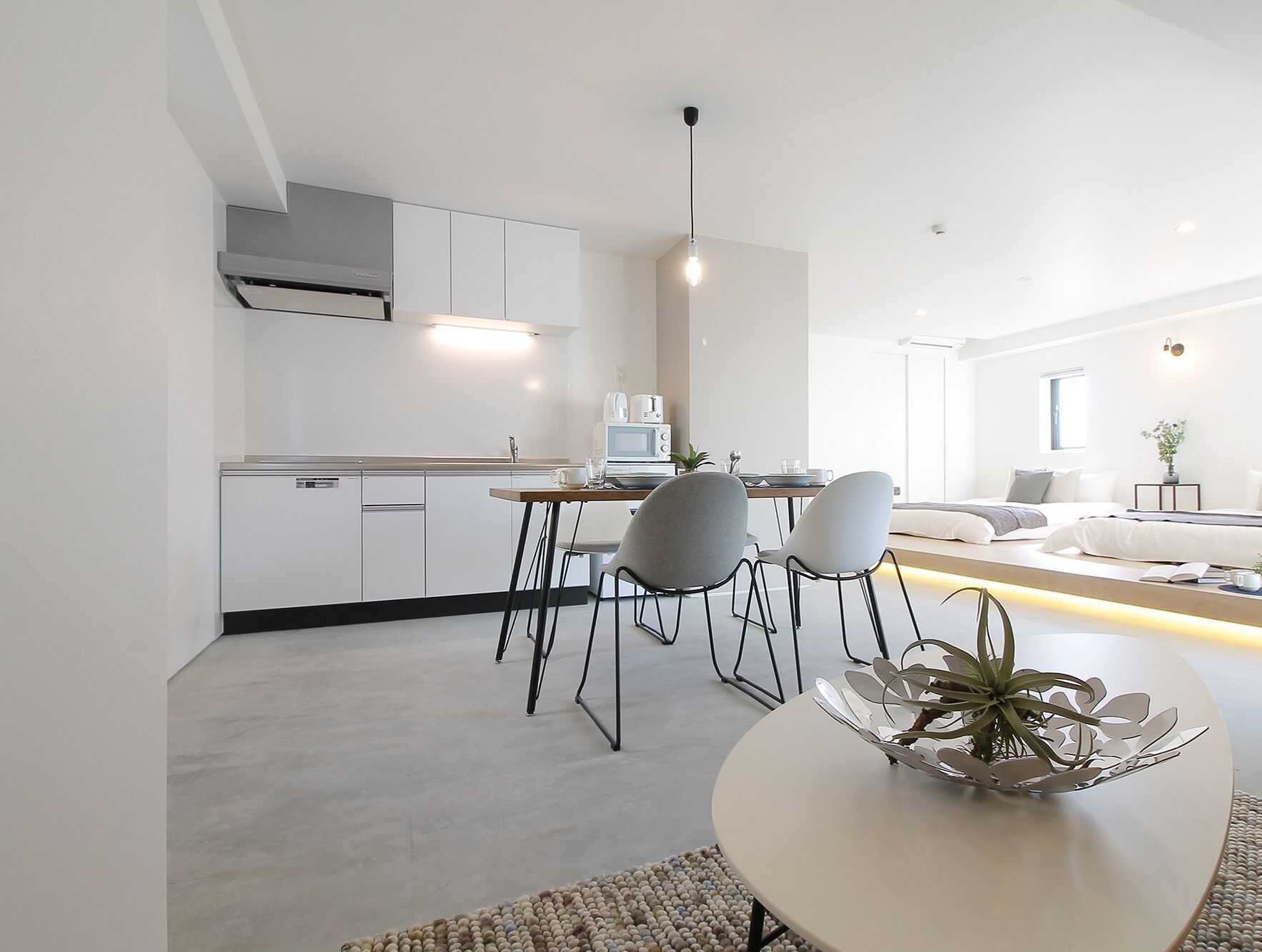 Una cocina moderna con armarios blancos, un comedor con mesa redonda y sillas, y una sala de estar con sofá y planta.