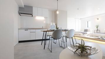 A modern kitchen with white cabinets, a dining area with a round table and chairs, and a living space with a sofa and a plant.