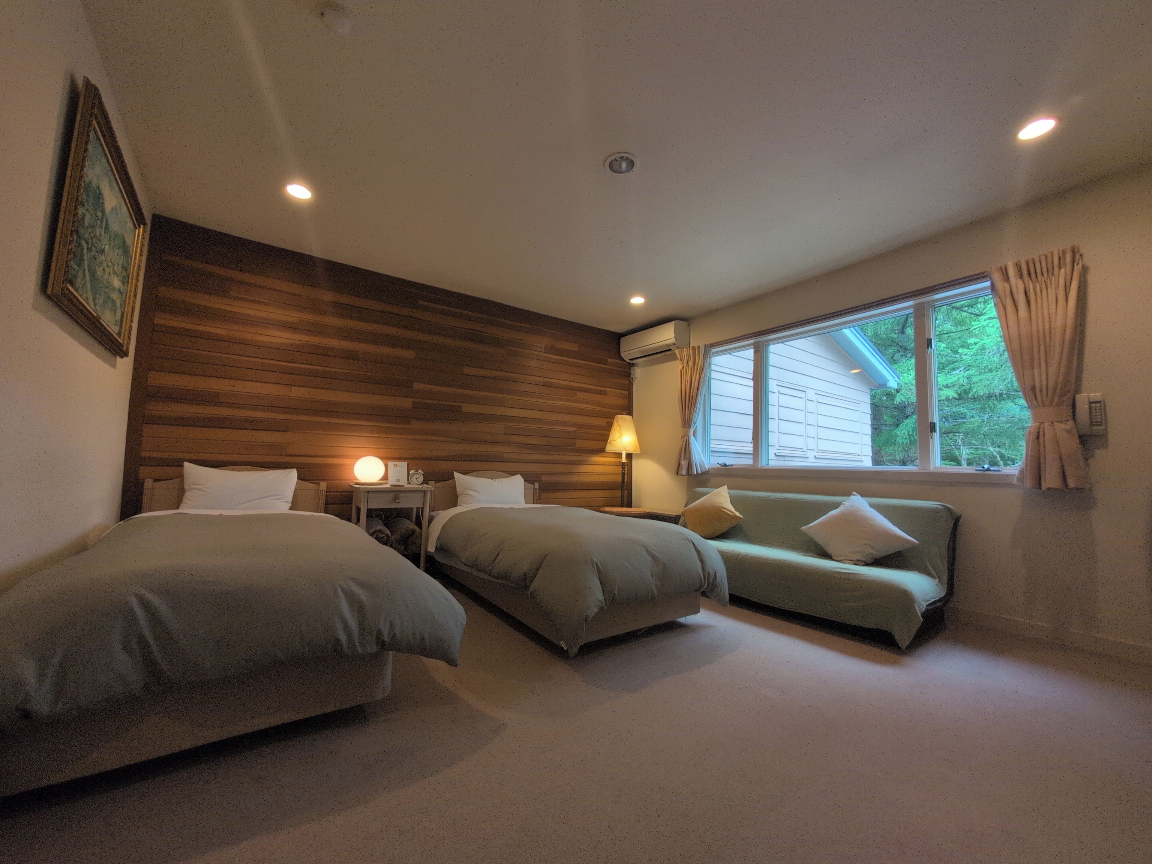 Une chambre d’hôtel avec deux lits, un canapé, une lampe, un appareil de climatisation et une fenêtre avec des rideaux.